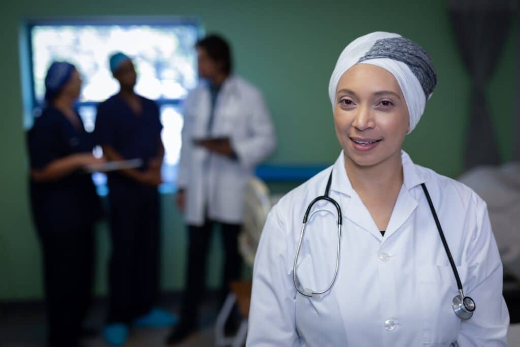 Front view of matured mixed race female doctor smiling into the camera while colleagues are discussing patient in the background in clinic at hospital